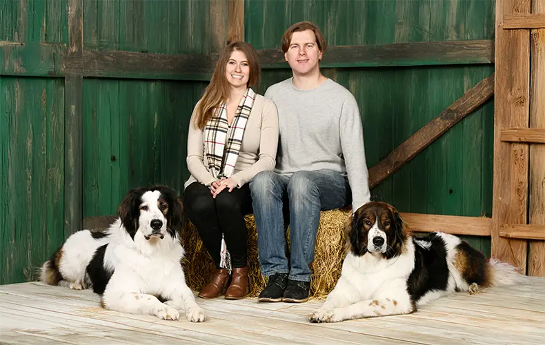 Owners of Od Dinare Kennel, Karina and Max, sitting on a hay bail in a barn with two imposing Tornjaks laying on either side of them